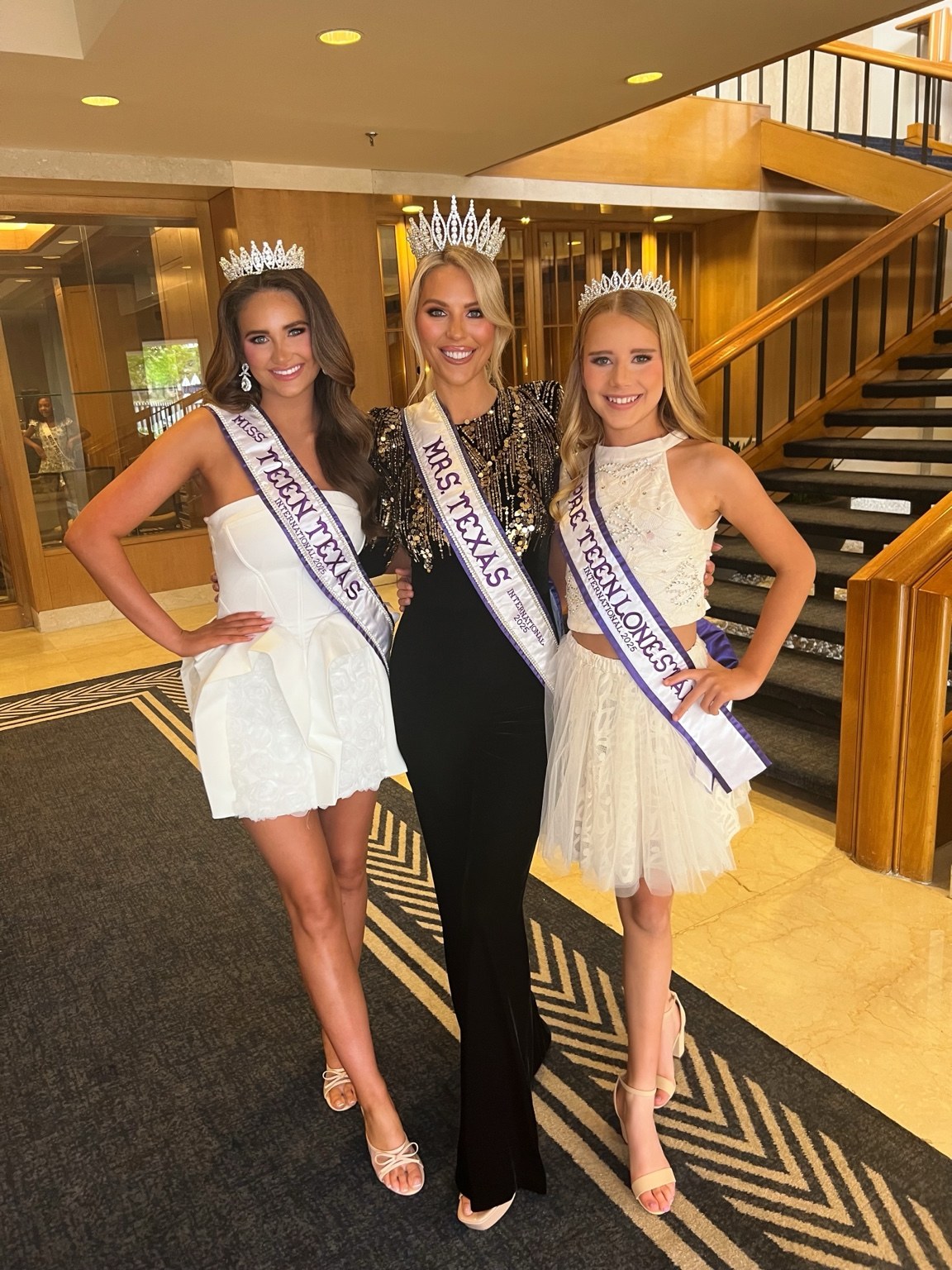 Three Texas International titleholders wearing crowns and sashes, posing together by a staircase